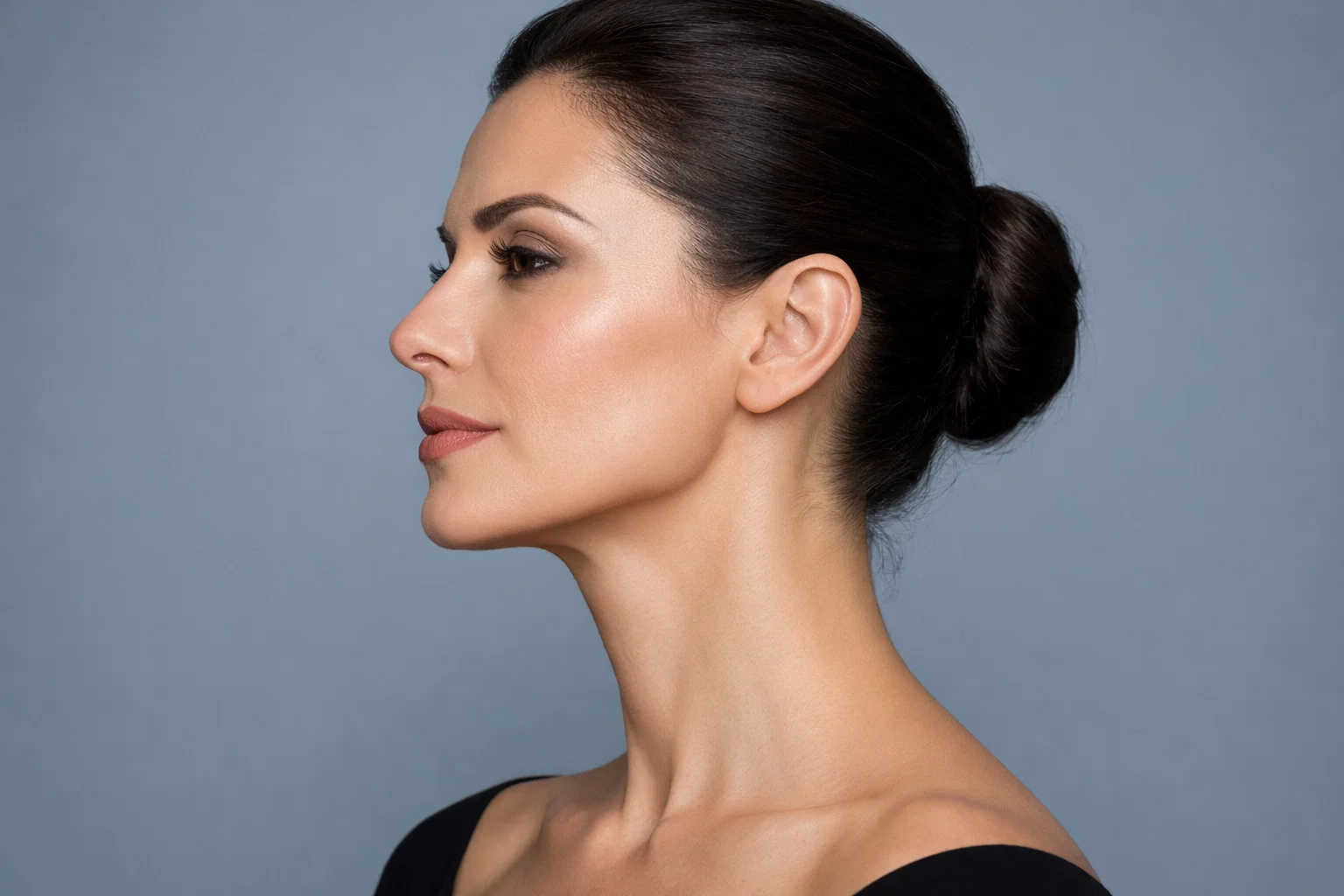 Side profile of a woman with a sculpted jawline and firm neck against a cool grey background.